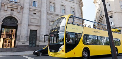 Żółty autobus piętrowy jedzie ulicą obok historycznego budynku.
