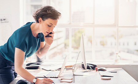 Kobieta rozmawiająca przez telefon i robiąca notatki w słonecznym biurze z dużymi oknami
