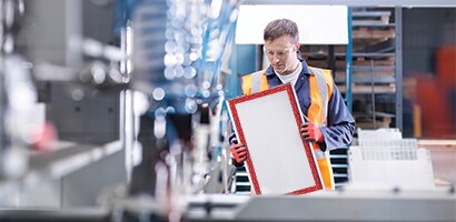 Pracownik w kamizelce ochronnej trzyma prostokątny panel na hali produkcyjnej.
